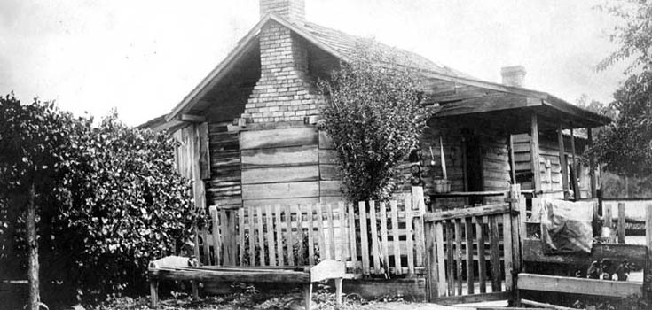 Black and white photo of wooden cabin with covered porch and brick chimney
