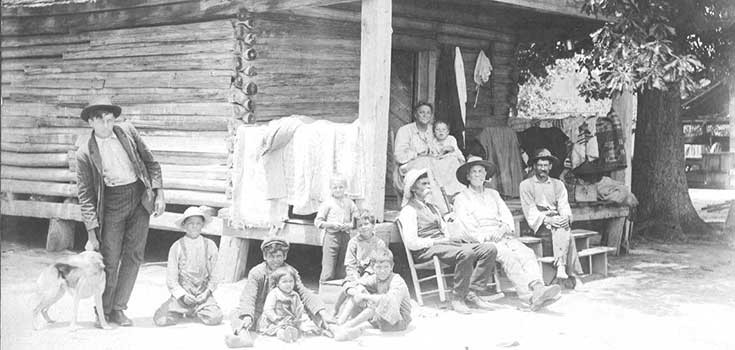 Black and white photo of a few adults and many small children in front of a small wood cabin